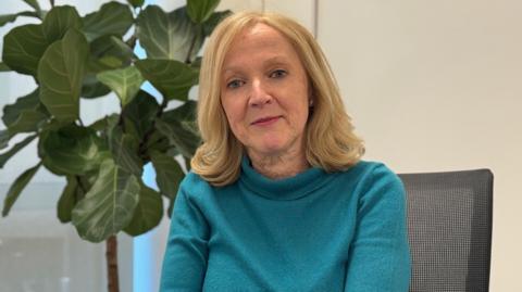 Helena Mangan, Head of Work and Family at Employment and Social Security for the States of Jersey wearing a teal long‑sleeved top sits in an office chair next to a large potted plant. The setting appears to be a bright indoor office space with light‑coloured walls and a window in the background.