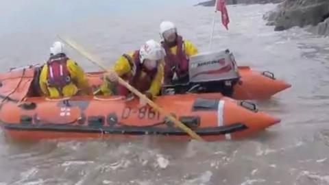 Three lifeboat crew members in yellow, all-in-one uniforms and white helmets, in a red dingy.