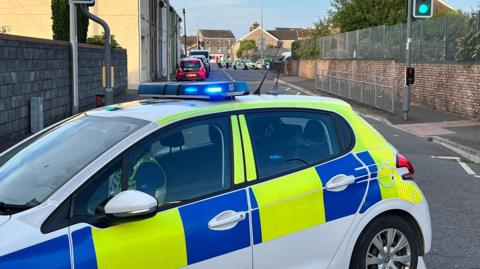 A police car in the forefront, with a road behind and, in the background at the far end of the road, tow more police cars and two uniformed officers.