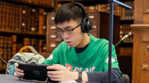  Marcus Koh is sitting at a table in a library holding a device. He has short, dark hair and is wearing spectacles and headphones. He is wearing a green top.