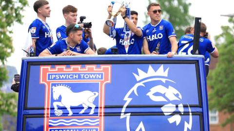 Ipswich Town players in an open-top bus with club branding are wearing their blue shirts and holding the Championship runners-up trophy aloft.