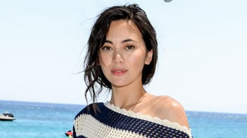 Jessica Henwick pictured in front of a blue ocean and a sunny, cloudless blue sky. She wears an off-the-shoulder, crocheted, black and white top. Her hair looks lightly windswept by the sea breeze.