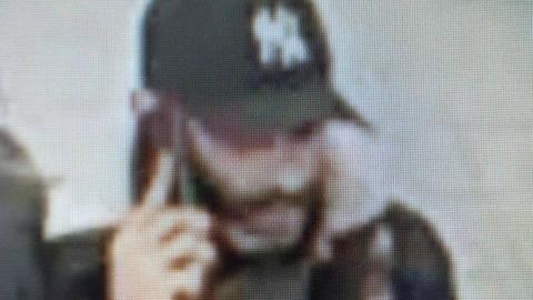 A blurry image of a man with brown facial hair with his right hand holding a mobile phone to his ear. He is wearing a black baseball cap with a white logo on the front