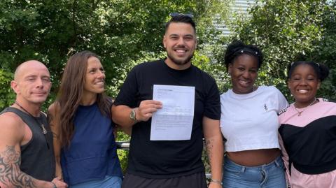 A smiling man in a black T-shirt holds up a GCSE certificate, surrounded by two teenage girls, a man and a woman.