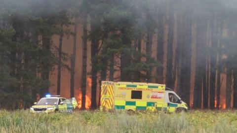 Flames are clearly visible in a forest. In the foreground is an ambulance and a paramedic car.