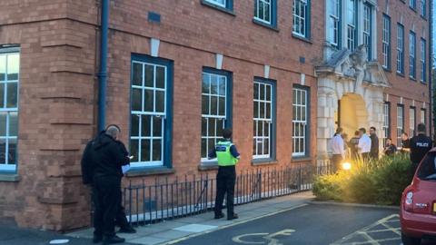 At least half a dozen people are standing outside an entrance to the building, which is on the right of the photo. Three other people are in the middle and left of the image.