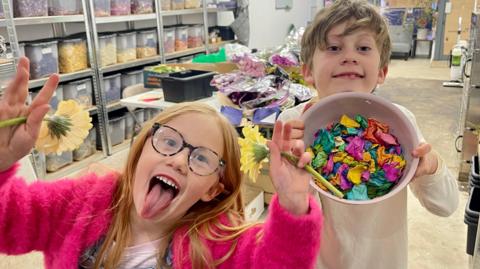 A girl and boy stand in a shop unit. The girl on the left has long red hair, glasses, a pink fluffy cardigan, a pink t-shirt and denim dungarees. She is holding a flower in each of her raised hands and is sticking her tongue out. The boy has short fair hair, a long sleeved white top and is holding a bowl of bright confetti. Behind them are trestle tables covered in bunches and boxes of flowers. There are rows of shelves with labelled transparent boxes full of different coloured flower petals. The floor is bare concrete and the shop-front windows are covered with cardboard. 