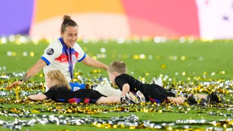 Lucy Bronze, who has brown tied-back hair, sat on the pitch after England's win. Her niece and nephew have dived into the confetti on the pitch next to her. She has her arms out wide and looks very happy.