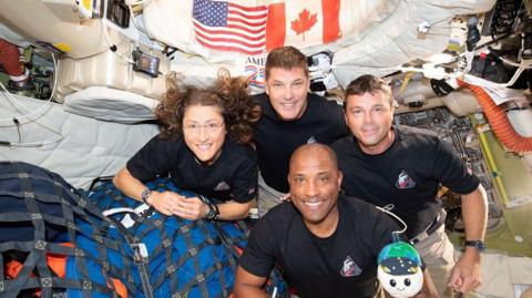 Four Artemis II crew members are inside the spacecraft, wearing matching dark shirts, with US and Canadian flags visible on the wall behind them. Left to right, they are Mission Specialist Christina Koch, Mission Specialist Jeremy Hansen, Commander Reid Wiseman, with Pilot Victor Glover below.
