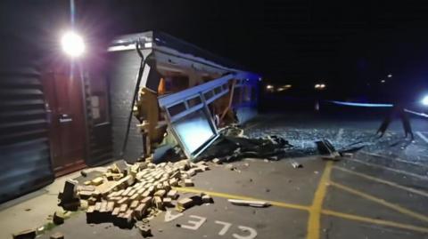 A black, single-storey building at the side of a tarmacked area, at night. There is broken glass and several bricks strewn across the tarmac and a big hole in the side of the building. White guttering and windows are hanging off the damaged building facade.