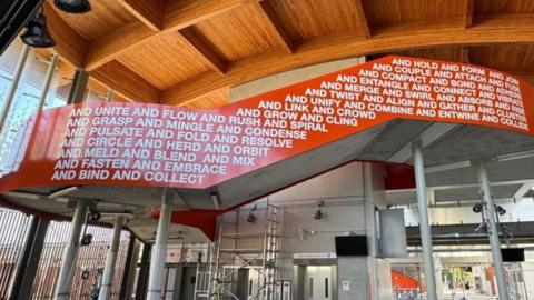 The interior of a new railway station, which still has some scaffolding up as it is not yet complete. The ceiling is wooden and beneath is a stairway balustrade, sweeping from left to right, which is coloured bright orange and has words written in white on it. 