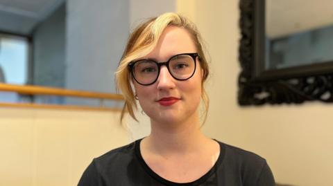 A white woman looking into the camera. She has black glasses and an eyebrow piercing. She is wearing red lipstick and a a black scoop neck T-shirt. Her blonde hair is tied up with a side parting.