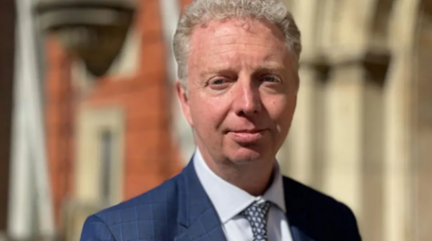 Alex Allinson is looking into the camera. He has short grey hair and is wearing a navy jacket and white shirt, and a tie.