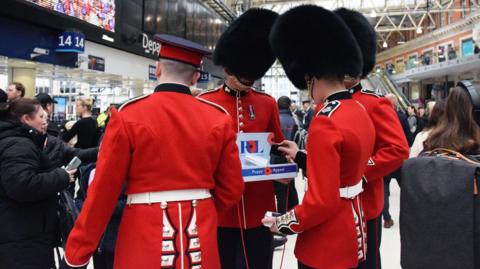 Grenadier Guards collecting at Waterloo on London Poppy Day. Four of the guards are standing in the busy train station. 