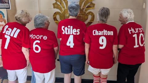Five women - all dressed in red England football shirts, standing in a line with their backs to the camera.