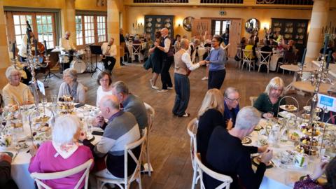 Groups of older people sit around round dining tables as they enjoy afternoon tea in a large dining room as people swing dance in the centre of the room
