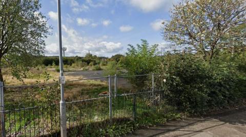 Site of the former recreation centre. The picture shows an area of grass which is yellow and green and a large patch of concrete behind railings and a lamp post. In front of it is a pavement. 