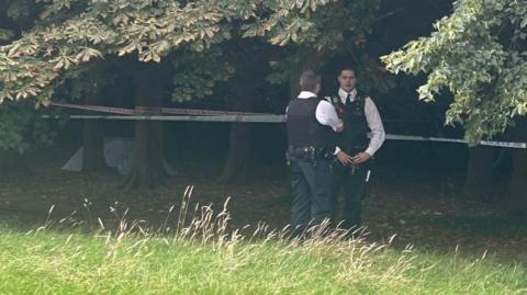 Image of two officers talking in front of a cordon around tree trunks. A small white tent can be seen in the distance