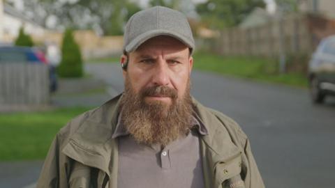 A man in a green jacket and grey shirt is standing in the middle of a road in a housing cul-de-sac. He has dark eyes and a long dark beard, which is going grey at the edges. The man is also wearing a grey cap and an earpiece. Behind him are trees, several small urban gardens and driveways, and cars parked at either side of him. 