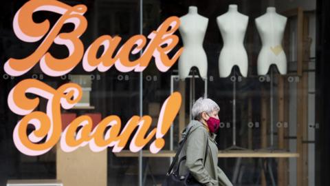 Woman in mask walks past closed shop with the words 'Back Soon!' on the window