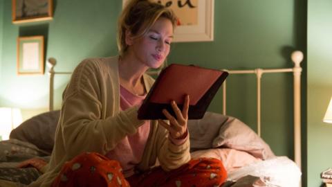 A still from the previous film, in which Bridget Jones sits on a bed in her pyjamas, holding an ipad.