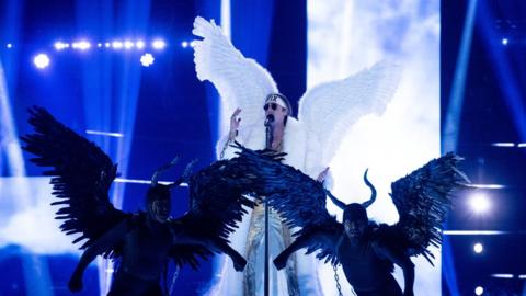 TIX on stage singing into the microphone dressed as an angel with sunglasses on, he is chained to two dancers dressed as winged demons who are looking into camera.