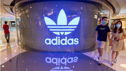 Shoppers wearing face masks walk past an Adidas logo at a shopping mall.