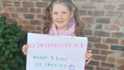 Charlotte holding a sign which says "#saveayricerink because I love skating"