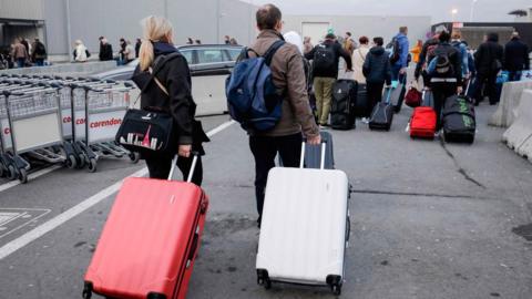 People are seen from from behind filing away in large queues, dragging suitcases behind them over the tarmac of the airport's roads