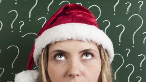 A woman wearing a Santa hat looks upwards, confused, set against a green chalkboard wall covered in chalk-written question marks