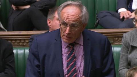 Man with grey hair and glasses, wearing a striped tie, speaks in the House of Commons
