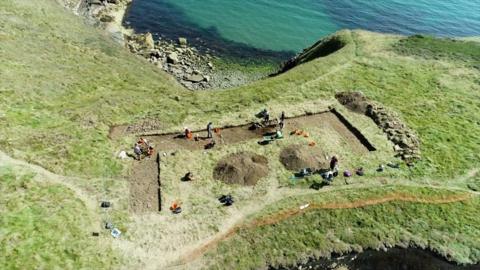 Aerial view of a hillfort on a peninsula with archaeologist working on a square dig