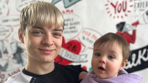 A photo of young dad Tyler holding his 10-month-old daughter Aurianna. Both are smiling. Tyler has dyed blonde hair and his long fringe is down to his eyebrows. He has a small black nose stud in and is wearing a black and white T-shirt. Aurianna has brunette hair and is wearing a purple jumper. It's a close up photo of their heads and shoulders and behind them is a mural on the wall with positive phrases on about being a dad.