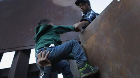 A boy is hoisted by fellow members of the migrant caravan over the US-Mexico border fence