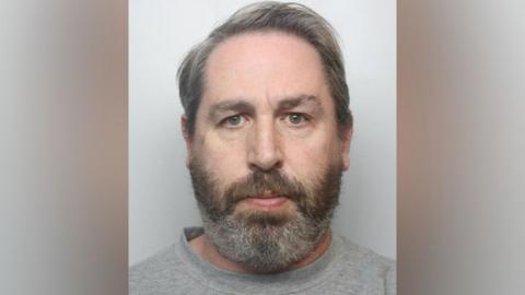 A mug shot of a grey haired man with a brown and white beard in grey clothing.