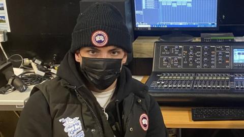 JM in the music studio, he's in a body warmer with a sticker on the breast which reads RIP Harvey and Kyres. Forever Young. He's wearing a wooly hat and a black face mask.