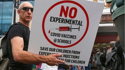 man holding sign reading 'no experimental covid vaccines @schools'