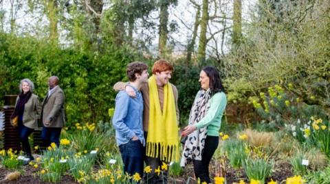 Three people standing among daffodils, chatting and laughing, with a couple in the background walking past the daffodils.