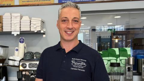 Daniele Panunzio wearing a black Panunzio's Kitchen branded polo shirt and smiling at the camera. He is standing behind the counter of his ice cream shop in Bristol, which is painted a pale blue and has shelves stacked with boxes, cups and coffee. In the foreground there is a glass jar full of baked treats and Mr Panunzio is holding a metal scoop of a pink gelato and posing for a picture.