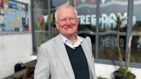 Steve Lansdown, a man with a grey blazer, blue jumper and white collared shirt smiling towards the camera. He has wispy grey hair. 