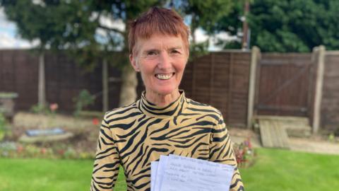 Tizi Hodson stands in the garden of her Lincolnshire home. She has short brown hair, is wearing a leopard-print top and is holding pages of a letter.