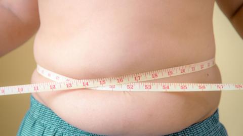 A topless child with blue shorts on with a tape measure around their waist. They are overweight.
