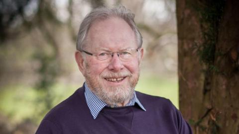 A man with grey hair, glasses and a grey beard, and wearing a purple jumper over a blue and white-collared shirt, smiling at the camera.