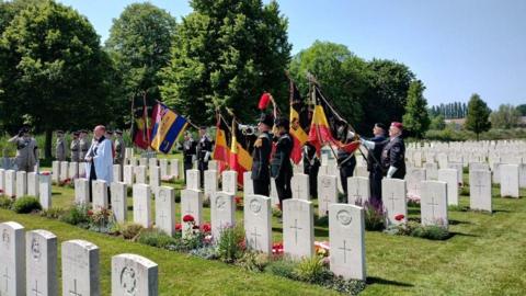 A padre wearing a white cloak and men in military uniform holding up flags, standing in a cemetery with neat rows of white graves. It's a sunny day and the sky is blue.