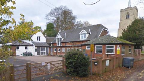 A primary school with a yard in to the left and old two-storey brick buildings to the right. There is a white building with a triangular upper floor in the background, and a brick wall in the foreground with a wooden gate across the entrance. There is a church to the right.