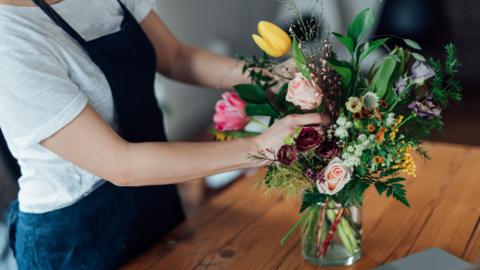 Person in a white shirt and dark blue apron arranging a colourful bouquet of tulips, roses, lisianthus, and foliage in a glass vase on a wooden table