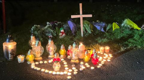 A cross has been placed on grass. Several candles are lit. It's night. Tea candles are in the shape of a heart.