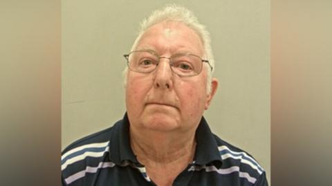 A mugshot of a man in a striped polo shirt. He is wearing glasses and has grey hair. 
