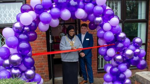 The foreground shows a large balloon arch made up of balloons in different shades of purple.  Inside the arch is  Lord Mayor of Sheffield Councillor Safiya Saeed cutting a red ribbon accompanied by Porter Brook Medical Centre Business and Network Manager Tom Hewitt. 
In the background is the redbrick building housing the expanded Porter Brook Medical Centre.