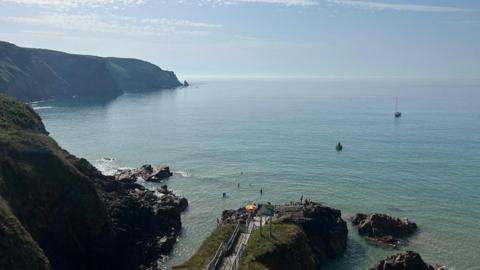 Steps lead down to the sea with headlands to the left. The sky is hazy and a small yacht can be seen offshore.